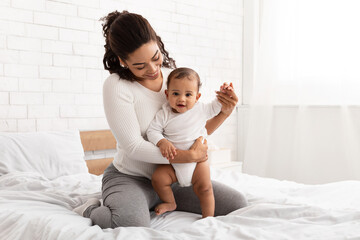 Black Mother Playing With Infant Baby Son Sitting In Bed