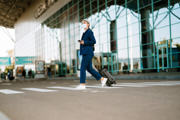 Fototapeta na wymiar Grey woman in face mask using cellphone while walking on zebra crossing