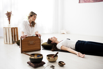 A yoga teacher gives a woman a sound bath lying on the mat. She plays a shruti box from India and there are several Tibetan bowls too