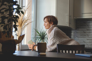 Beautiful middle-aged baby boomer woman in beige sweater holds phone and looks out window. Background of light gray interior.