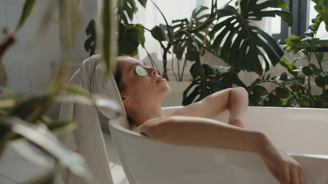 Pano shot of relaxed bautiful caucasian young woman lying in spa bath with cucumber on eyes and white towel on head, eyes closed. Relaxing spa wellness treatments in eco design bathroom