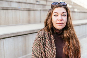 Female in checkered coat and turtleneck with long hair looking at camera while sitting on concrete stairs in daytime in city