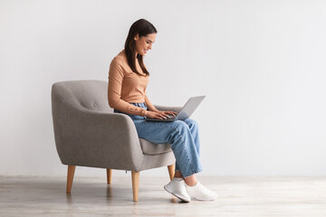 Happy young lady sitting in armchair, using laptop, studying remotely or having business meeting,...