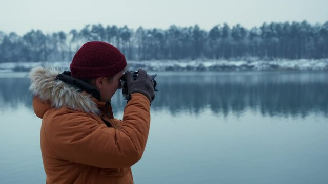 Traveler in an orange jacket takes a picture with a modern camera against the background of a winter landscape