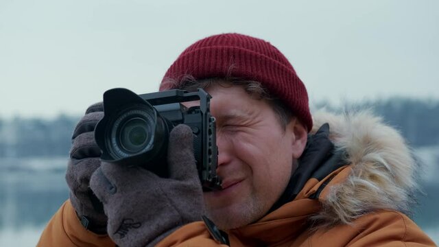 Travel man in an orange jacket and a red hat takes a picture with a modern camera against the background of a winter landscape.