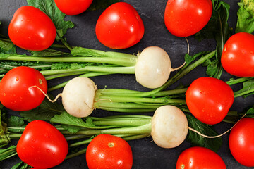 Vibrant red tomato and white radish roots with green leaves on dark gray table - view from above, healthy vegetables concept