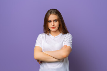 Fototapeta premium Portrait of upset brunette woman in white casual t shirt standing with arms folded over purple background. Attractive young woman in closed posture