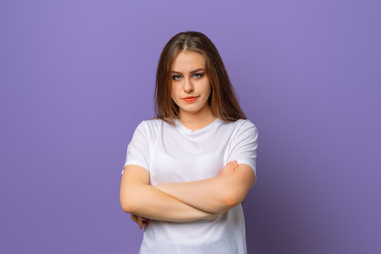 Portrait of upset brunette woman in white casual t shirt standing with arms folded over purple background. Attractive young woman in closed posture