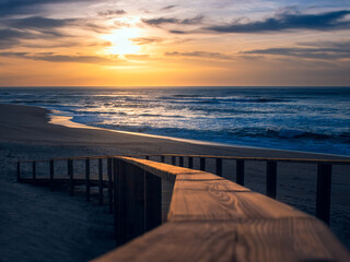 Landscape of golden sunset at the beach showing blurry close-up of illuminated wooden handrail of boardwalk leading down to the sand and Atlantic Ocean at Quiaios Beach, Portugal © Marta Nogueira