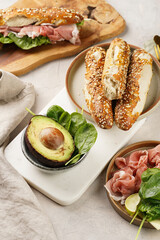 Savoury breakfast ingredients: brown lye bread sticks garnished with oats and seeds, pork slices prosciutto, hard cheese gouda and fresh spinach and avocado on marble board