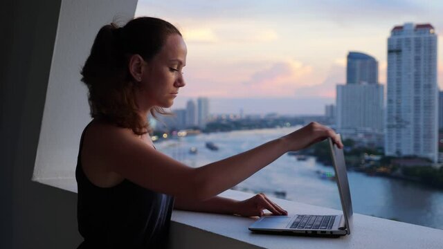 Woman finish work and close laptop, turn head and look down to beautiful evening city. Portrait shot of European freelancer against blurred Bangkok and Chao Phraya river view