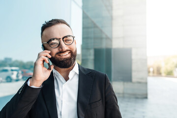 Happy CEO or manager talking on mobile phone and smiling while walking near office center during break, empty space