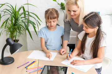 Woman tutor or foster parent mum helping cute caucasian school child girl doing homework sitting at table. Diverse nanny and kid learning writing in notebook studying at home