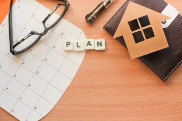 PLAN letterings on white cubes on the calendar. A house model on the notebook, pen, and eyeglasses. Business and plan concept