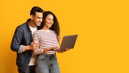 Young middle eastern couple using laptop together while standing over yellow background