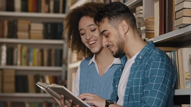 Young couple students standing in university library laughing reading funny story hispanic guy with african american girl holding book in hands studying looking at textbook school college education