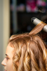 Fototapeta premium Hairdresser makes curls with a curling iron for the young woman with long brown hair in a beauty salon