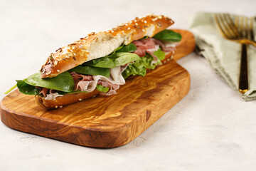Long sandwich in brown lye bread sticks garnished with oats , pork slices prosciutto, hard cheese gouda and fresh greens on wooden board, beige surface