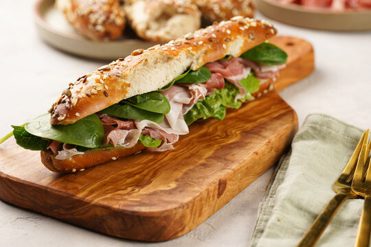 Long sandwich baguette in brown lye bread sticks garnished with oats , pork slices prosciutto, hard cheese gouda and fresh greens on wooden board, beige surface