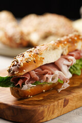 Long sandwich in brown lye bread sticks garnished with oats , pork slices prosciutto, hard cheese gouda and fresh greens on wooden board, beige surface