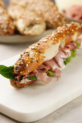 Long sandwich in brown lye bread sticks garnished with oats , pork slices prosciutto, hard cheese gouda and fresh greens on marble board, beige surface
