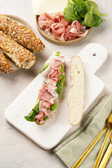 Long sandwich in brown lye bread sticks garnished with oats , pork slices prosciutto, hard cheese gouda and fresh greens on marble board, beige surface