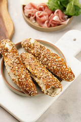Savoury breakfast ingredients: brown lye bread sticks garnished with oats and seeds, pork slices prosciutto, hard cheese gouda on marble board