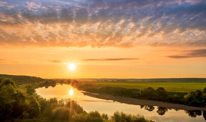Scenic view at beautiful sunset or sunrise on a shiny river with green bushes on sides, golden sun rays, calm water ,deep blue cloudy sky and forest on a background, spring landscape