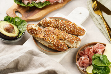 Savoury breakfast ingredients: brown lye bread sticks garnished with oats and seeds, pork slices prosciutto, hard cheese gouda and fresh spinach and avocado on marble board