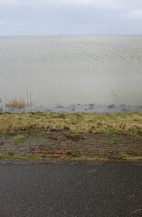 Edge of North Sea protection dyke with tarmac road and red brickstone bank during high tide on a rainy day (vertical image), Burhave, Lower Saxony, Germany