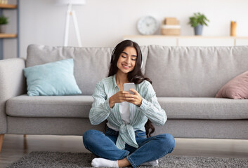Fototapeta premium Full length of millennial Indian lady in headphones using cellphone, listening to music, sitting on floor at home