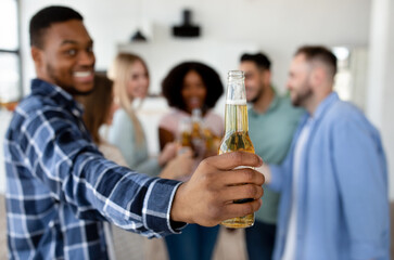 Funky black guy showing bottle of beer, celebrating with group of his diverse friends, saying CHEERS at home, closeup
