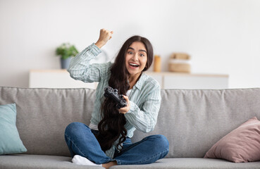 Fototapeta premium Full length fo excited Indian woman with joystick sitting on couch at home, making victory gesture, winning video game