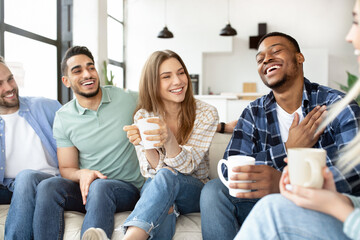 Group of diverse friends spending time togeher on weekend, holding hot drinks, having conversation and laughing at home