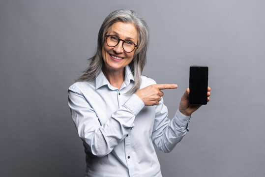 Mature gray-haired businesswoman wearing formal clothes showing smartphone and pointing with a finger on the screen with blank space for advertisement, standing isolated over gray background