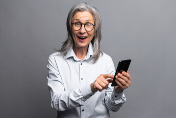 Surprised business woman in formal shirt pointing finger at smartphone while feeling shocked with new phone features. Indoor studio shot isolated on grey background
