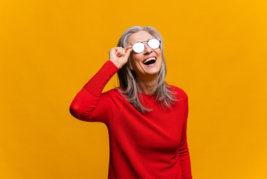 Charming senior woman in sunglasses laughing toothy. Elegant carefree mature modern gray-haired lady in red jumper standing and laughing out loud over yellow background. Happy lifestyle concept