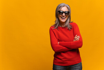 Merry senior middle-aged woman wearing sunglasses standing with arms crossed isolated on yellow....