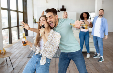 Millennial multiethnic couple singing song into microphone, drinking beer, having party with...