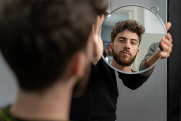 A young man looking at his new hair and freshly trimmed beard in the mirror held by the hairdresser
