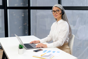 Cheerful aged woman with headset, wrinkles on the face, smiling, listening at the computer, consulting the client online , gives the advice, working, positive person