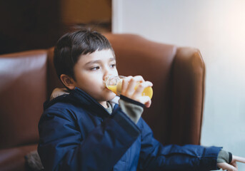 Healthy Kid boy drinking fresh orange juice in the cafe with bright light shining from window, Child sitting on sofa in coffee shop looking out with thinking face while drinking juice after snack.
