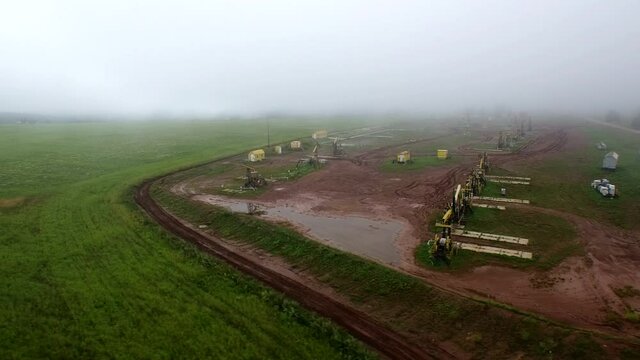 Aerial flying out view on oil pumps at oilfield cluster in a foggy field after rain. Muddy ground puddles hard working conditions.