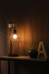 Stylish lamp, decor and green plant on wooden table near brown wall indoors. Interior design