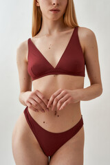 Fototapeta premium Cropped shot of slim young caucasian woman in burgundy underwear standing and posing isolated over light gray background