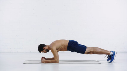 side view of shirtless sportsman doing plank exercise on fitness mat near white brick wall