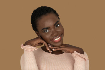 Closeup portrait of a smiling young black woman with short Afro hair, light makeup and lipstick posing by herself resting her head on the palm of her hand inside a studio with a pecan background.