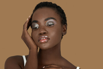 Closeup portrait of a serene young black woman with short Afro hair, light makeup and lipstick posing by herself inside a studio with a pecan background resting her palm of her hand on her face.