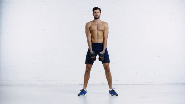 full length of sportive and shirtless man in shorts and sneakers standing with dumbbells against white brick wall