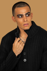 Closeup portrait of a serene young Latin man with short hair, light makeup and eyeshadow sitting by himself inside a studio with a pecan background wearing black jeans and a black sweater.